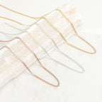 A variety of curb chain necklaces in gold, silver, and rose gold finishes displayed draped across a textured cream prop.