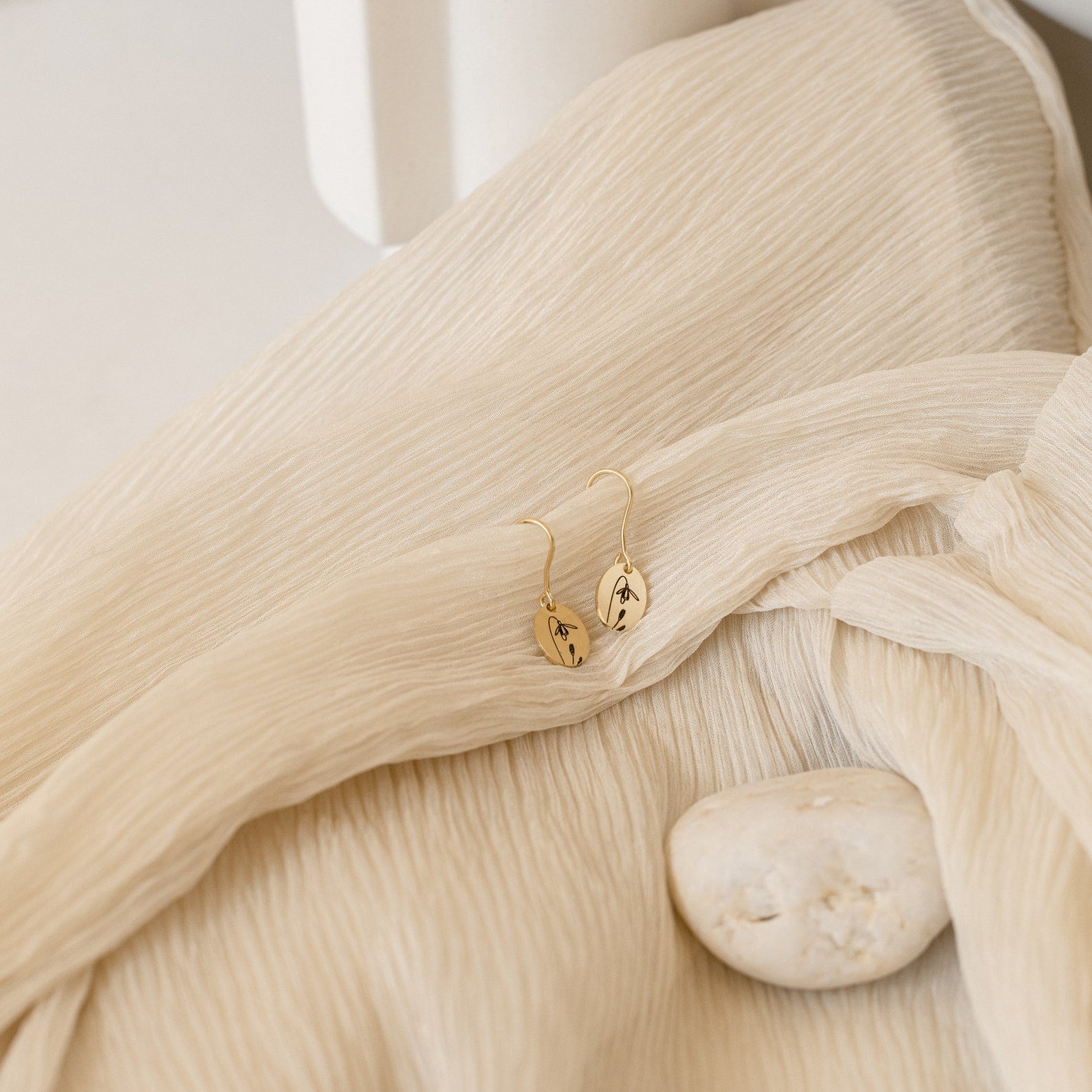Dangling Birth Flower Earrings rest on textured beige fabric next to a smooth white stone.
