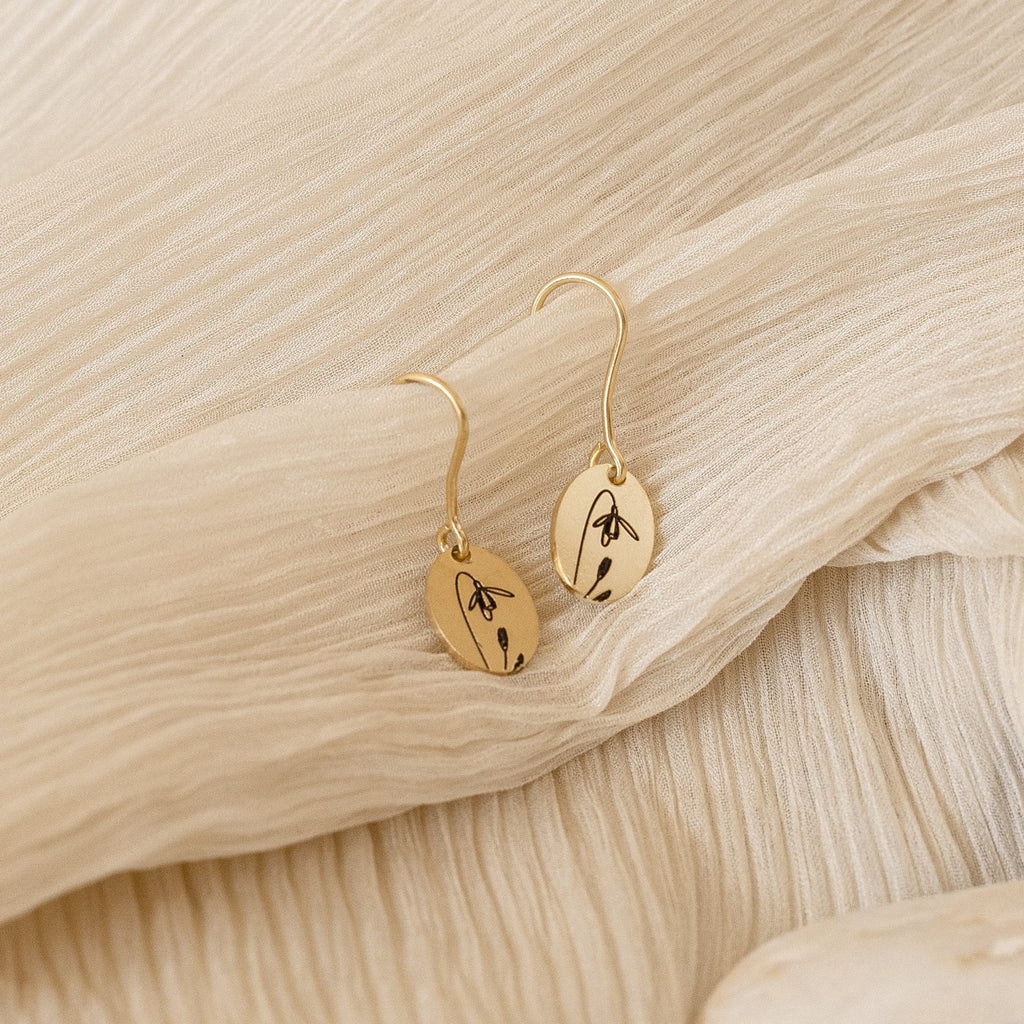 Dangling Birth Flower Earrings with gold floral engravings are displayed on textured cream fabric, adding a personalized touch to any outfit.