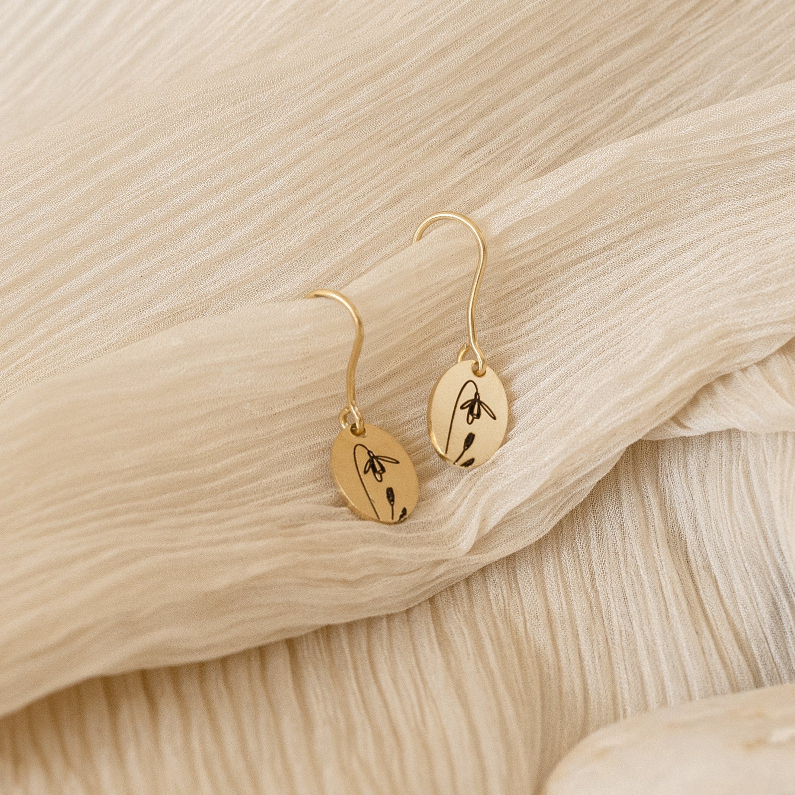 Dangling Birth Flower Earrings with gold floral engravings are displayed on textured cream fabric, adding a personalized touch to any outfit.