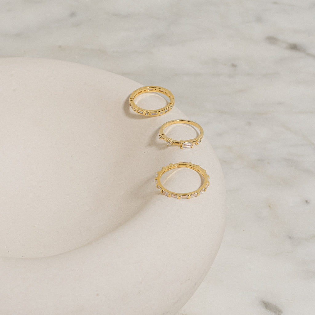 The Baguette Eternity Ring, featuring a delicate gold design, is displayed on a smooth white curved dish atop marble—a refined display of modern luxury.