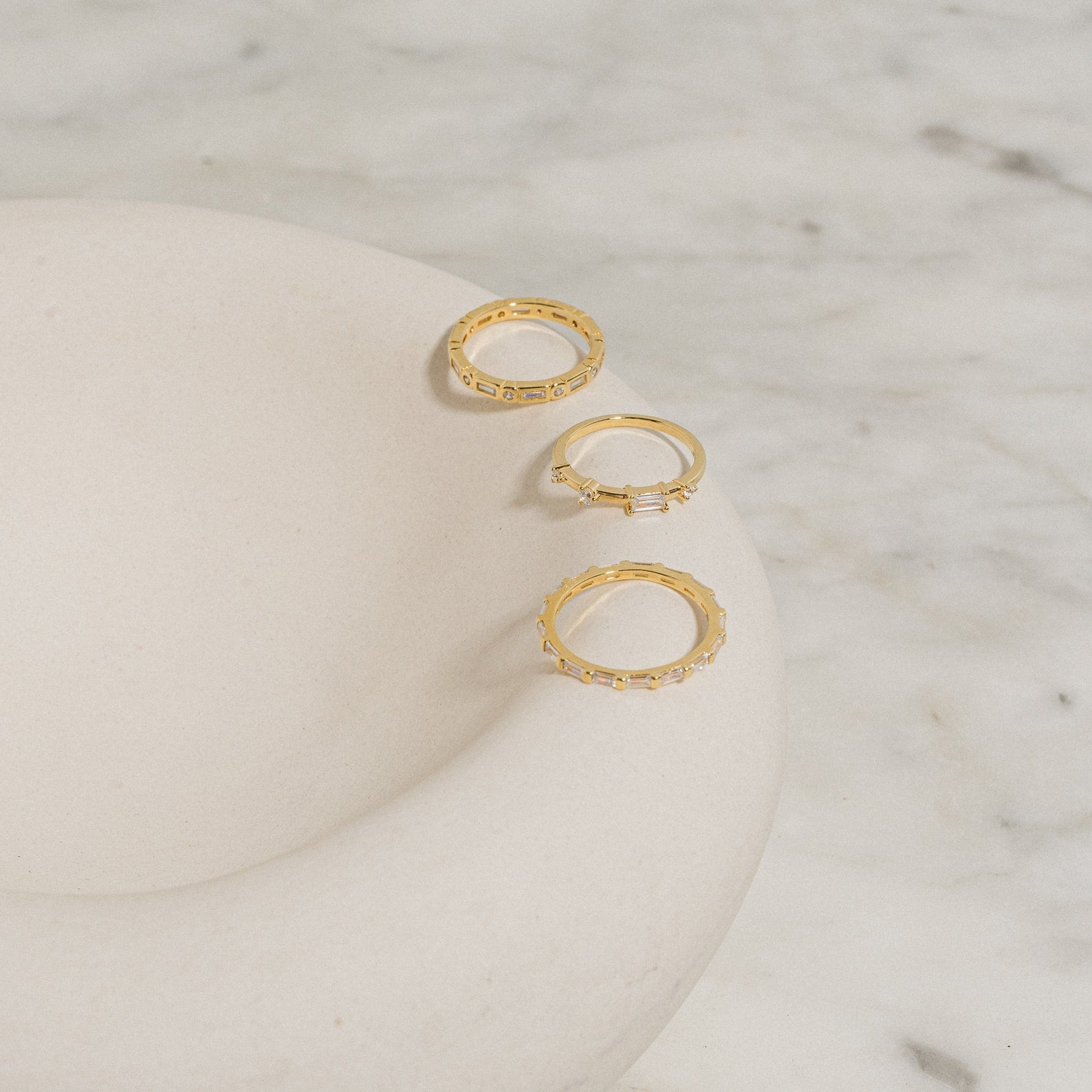 The Baguette Eternity Ring, featuring a delicate gold design, is displayed on a smooth white curved dish atop marble—a refined display of modern luxury.