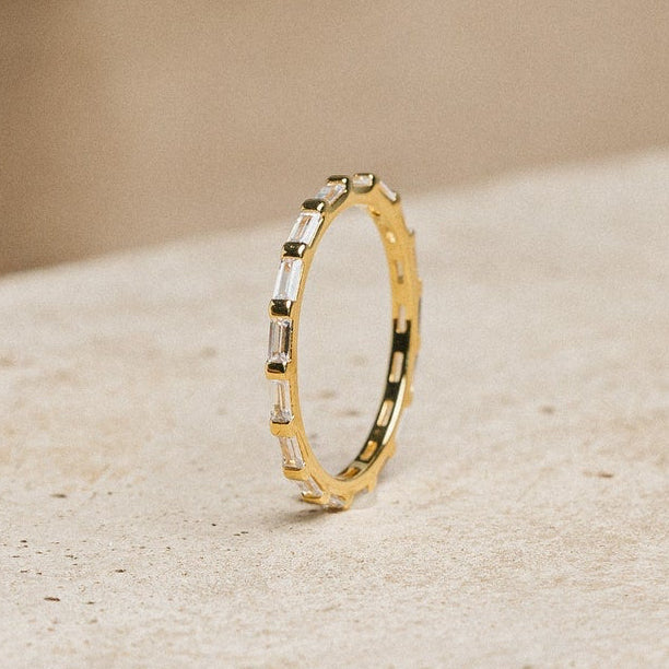 The Baguette Eternity Ring, featuring slim gold and rectangular baguette diamonds, is displayed on a textured concrete block with a beige backdrop, reflecting modern luxury.