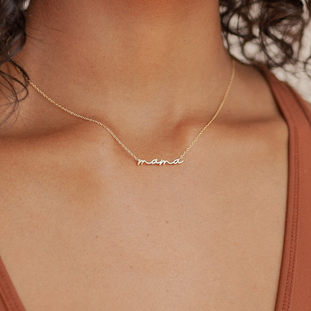Gold necklace with the word “mama” in smooth cursive script, worn just above the collarbone on a delicate chain. Styled with a rust colored V neck tank top for a minimal look.