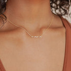 Gold necklace with the word “mama” in smooth cursive script, worn just above the collarbone on a delicate chain. Styled with a rust colored V neck tank top for a minimal look.