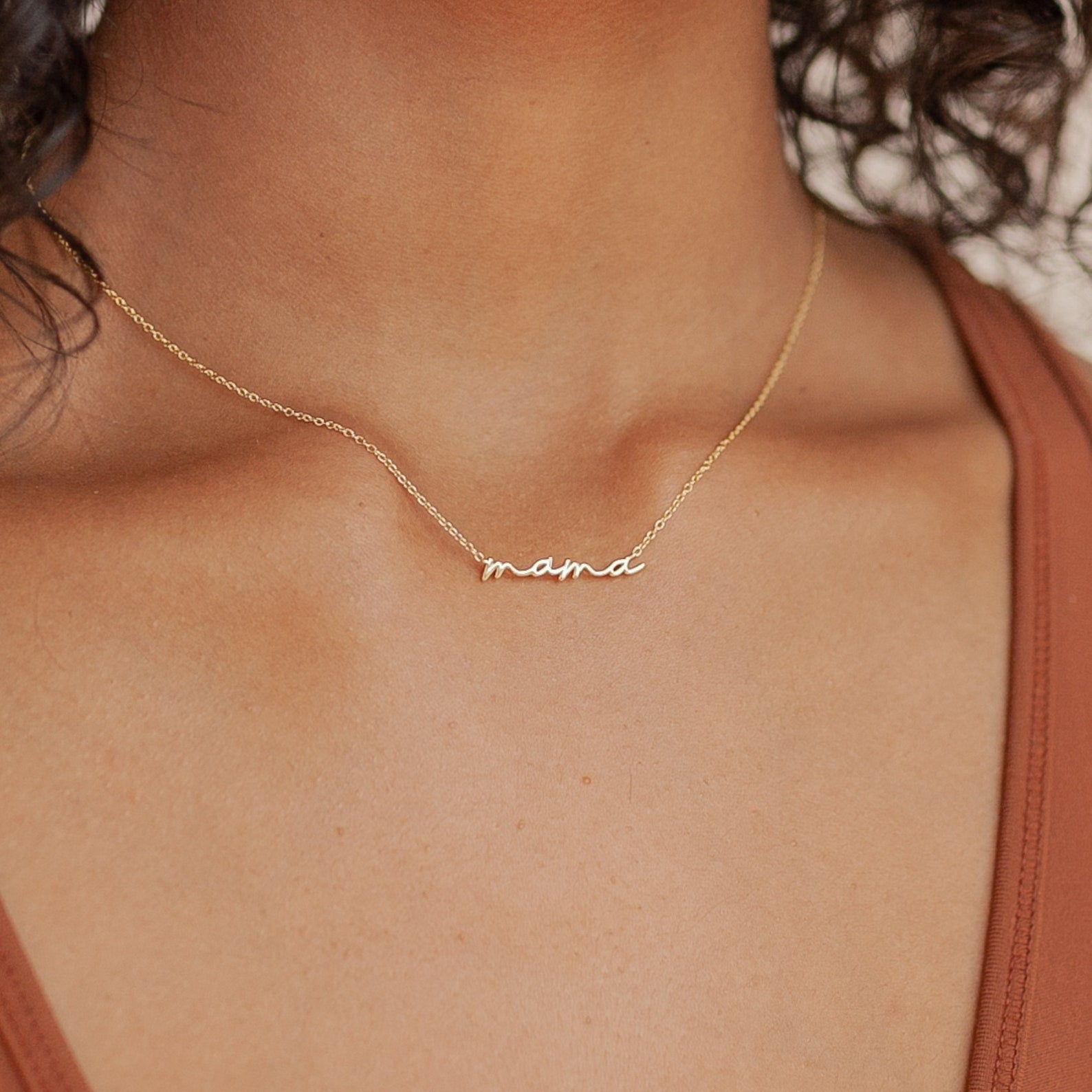 Gold necklace with the word “mama” in smooth cursive script, worn just above the collarbone on a delicate chain. Styled with a rust colored V neck tank top for a minimal look.