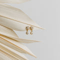Lexi Flower Studs, elegant gold and diamond earrings with a floral design, are displayed on dried palm leaves against a light gray background.