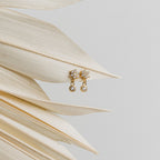 Lexi Flower Studs, elegant gold and diamond earrings with a floral design, are displayed on dried palm leaves against a light gray background.