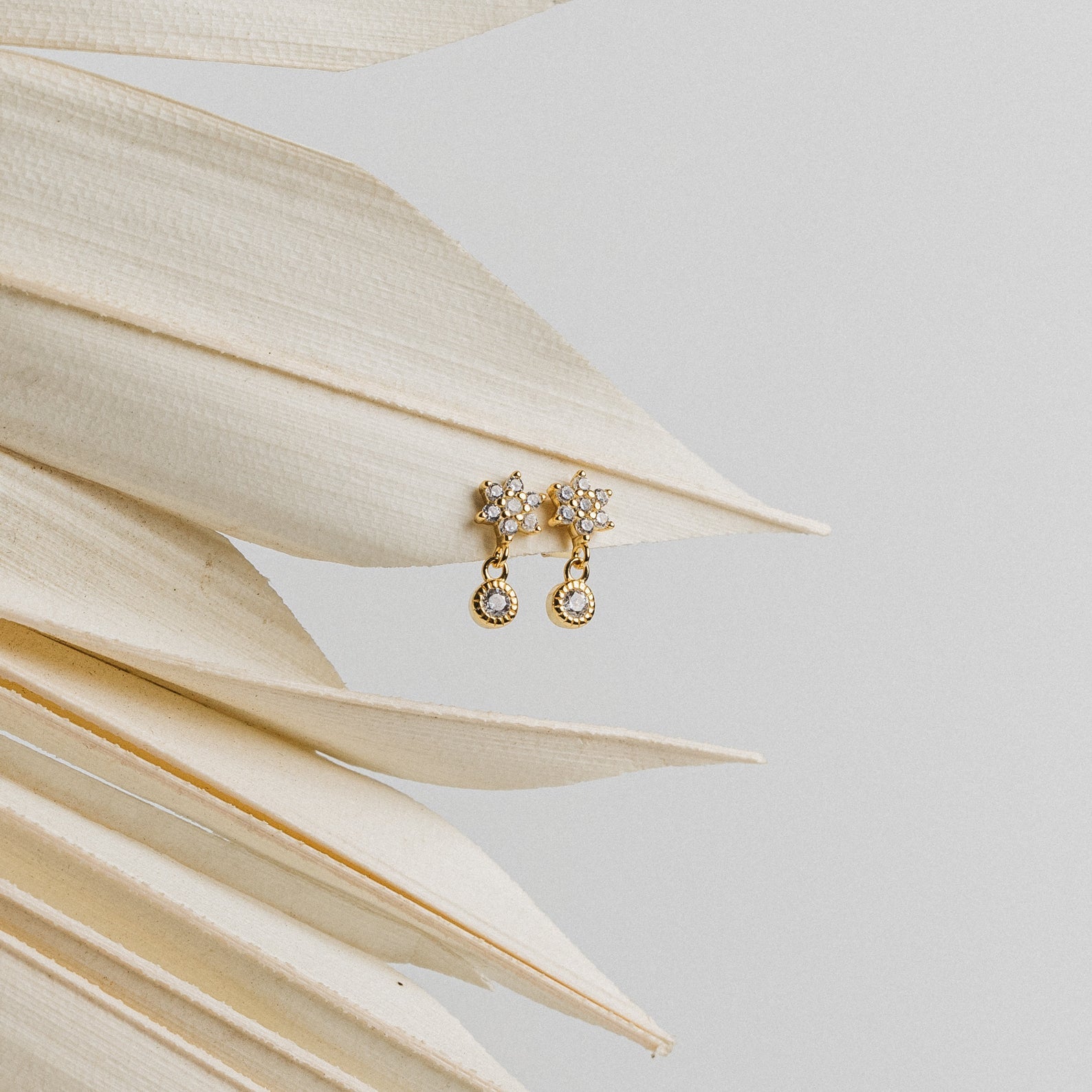 Lexi Flower Studs, elegant gold and diamond earrings with a floral design, are displayed on dried palm leaves against a light gray background.