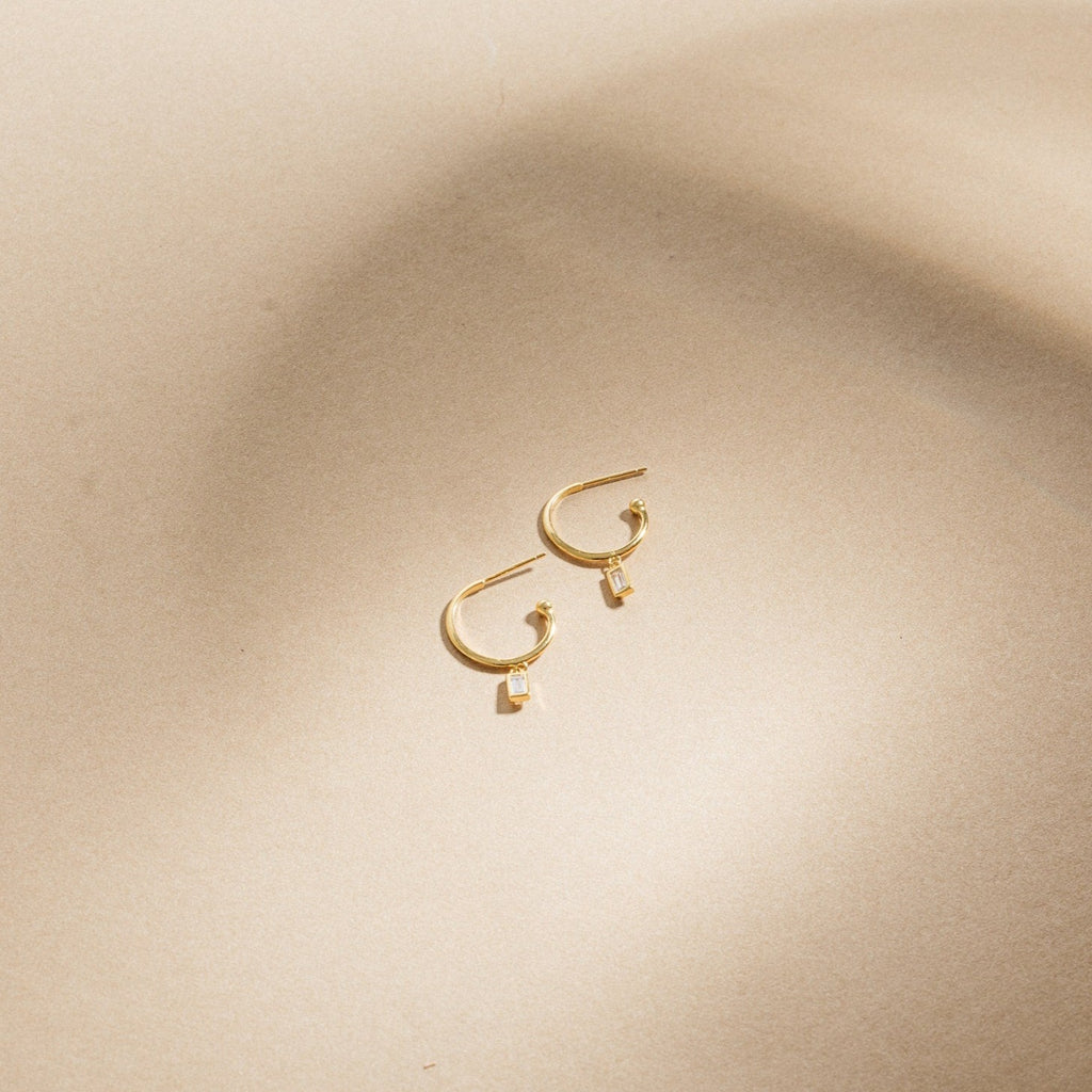 Elle Huggies—gold midi hoops with small rectangular pendants, inspired by the Elle Baguette Hoops—displayed on a smooth beige surface.