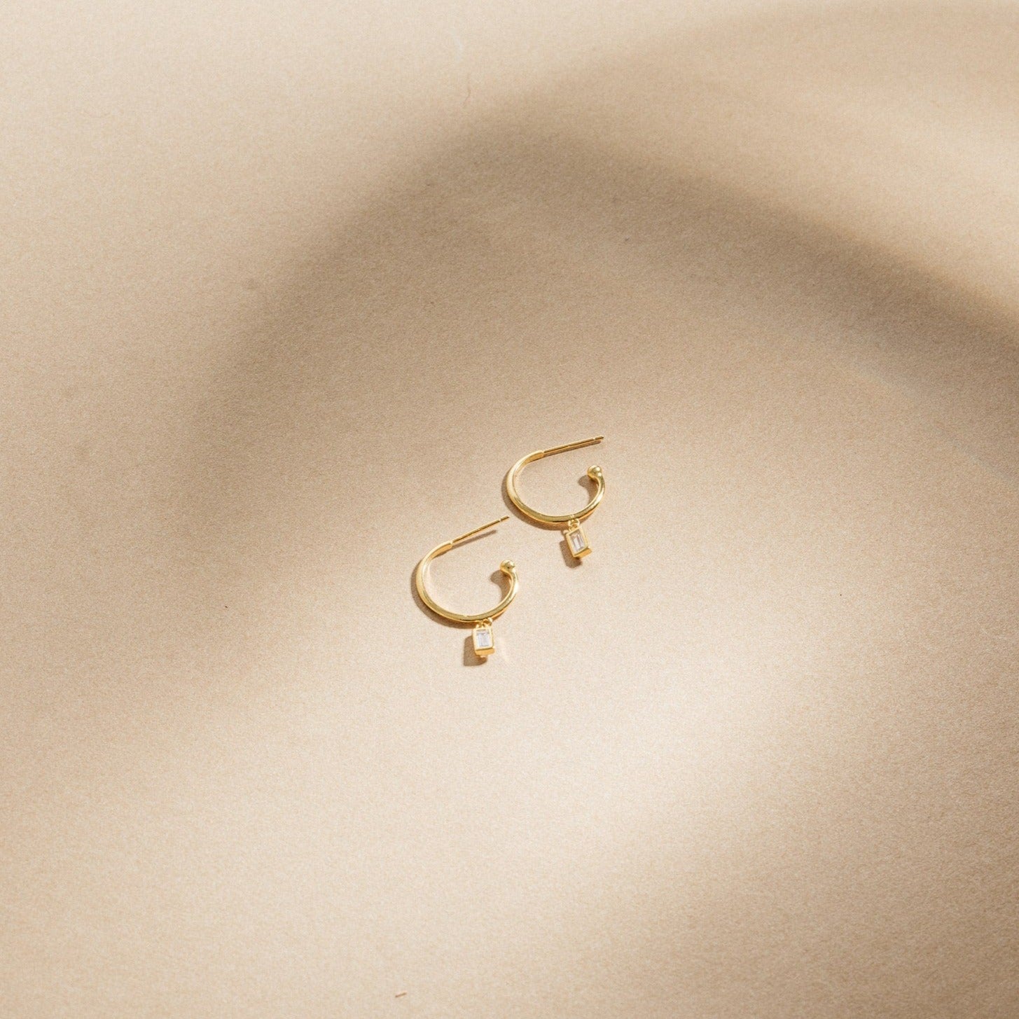 Elle Huggies—gold midi hoops with small rectangular pendants, inspired by the Elle Baguette Hoops—displayed on a smooth beige surface.