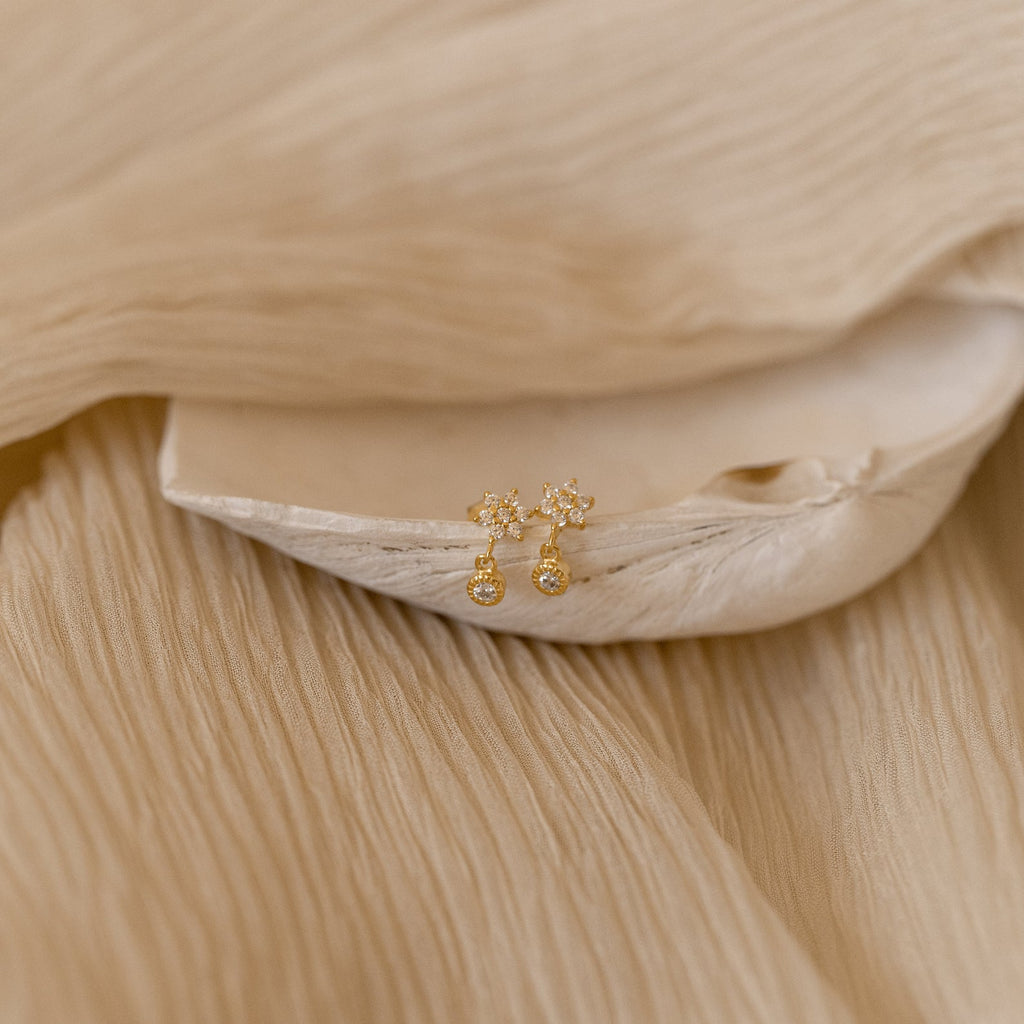 Lexi Flower Studs featuring gold floral designs with small clear stones, displayed on beige textured fabric and a shell.
