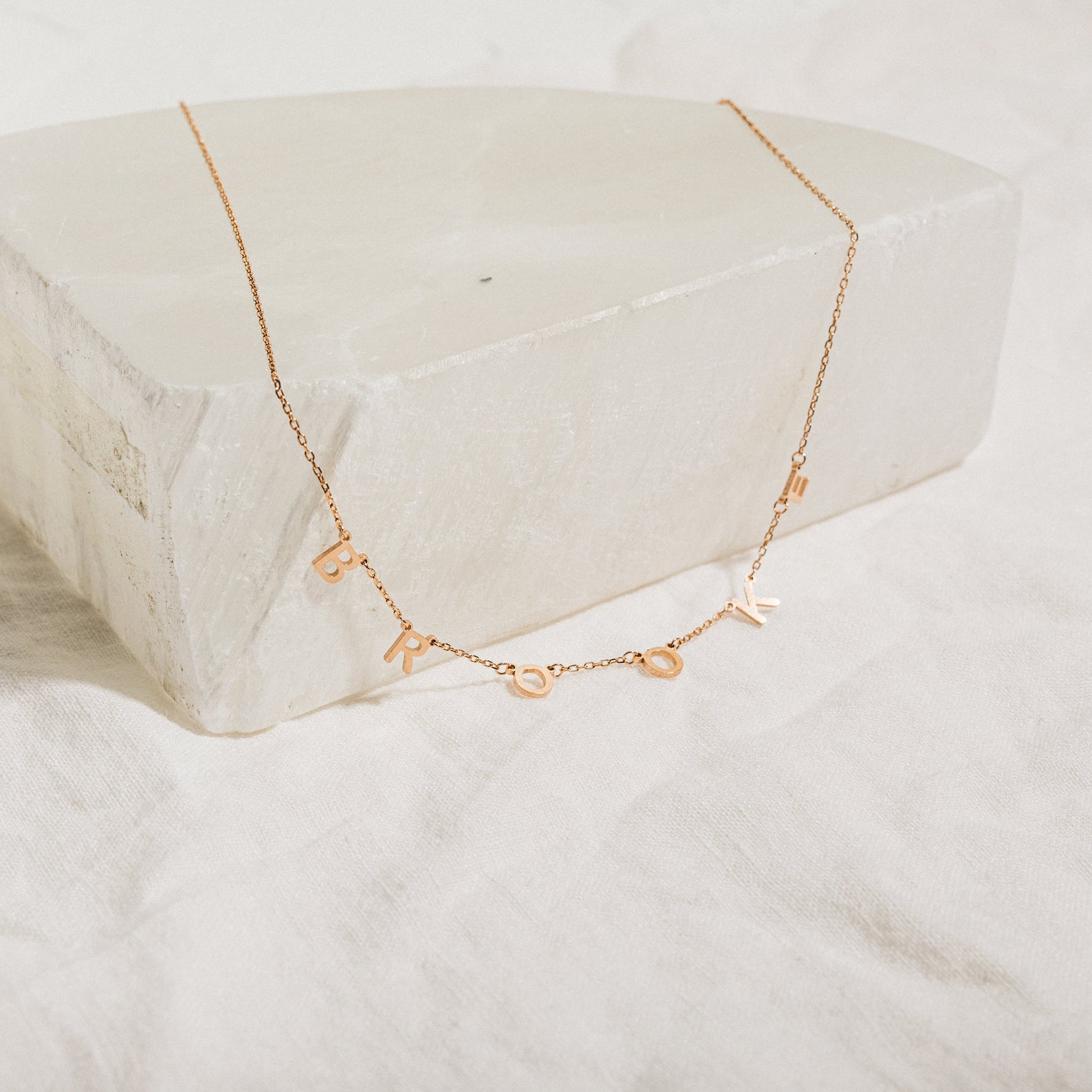 Custom Letter Necklace in Rose Gold featuring dangling charms that spell BROOKLYN, shown on a white stone block—a personalized necklace ideal for adding a unique touch.