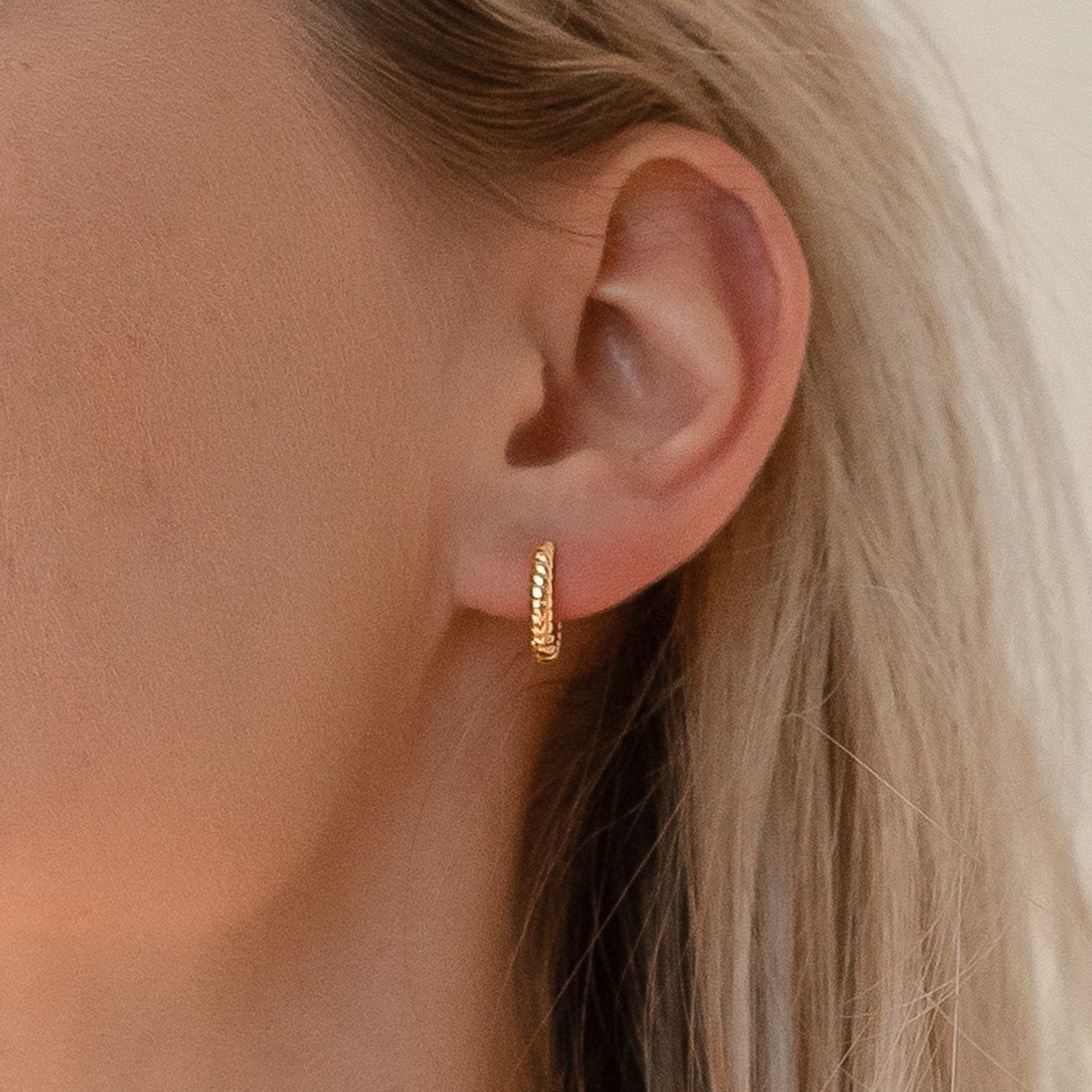 A close-up of a woman's ear wearing Thea Twisted Huggies, small gold earrings ideal for a minimalist look or an elegant ear stack.