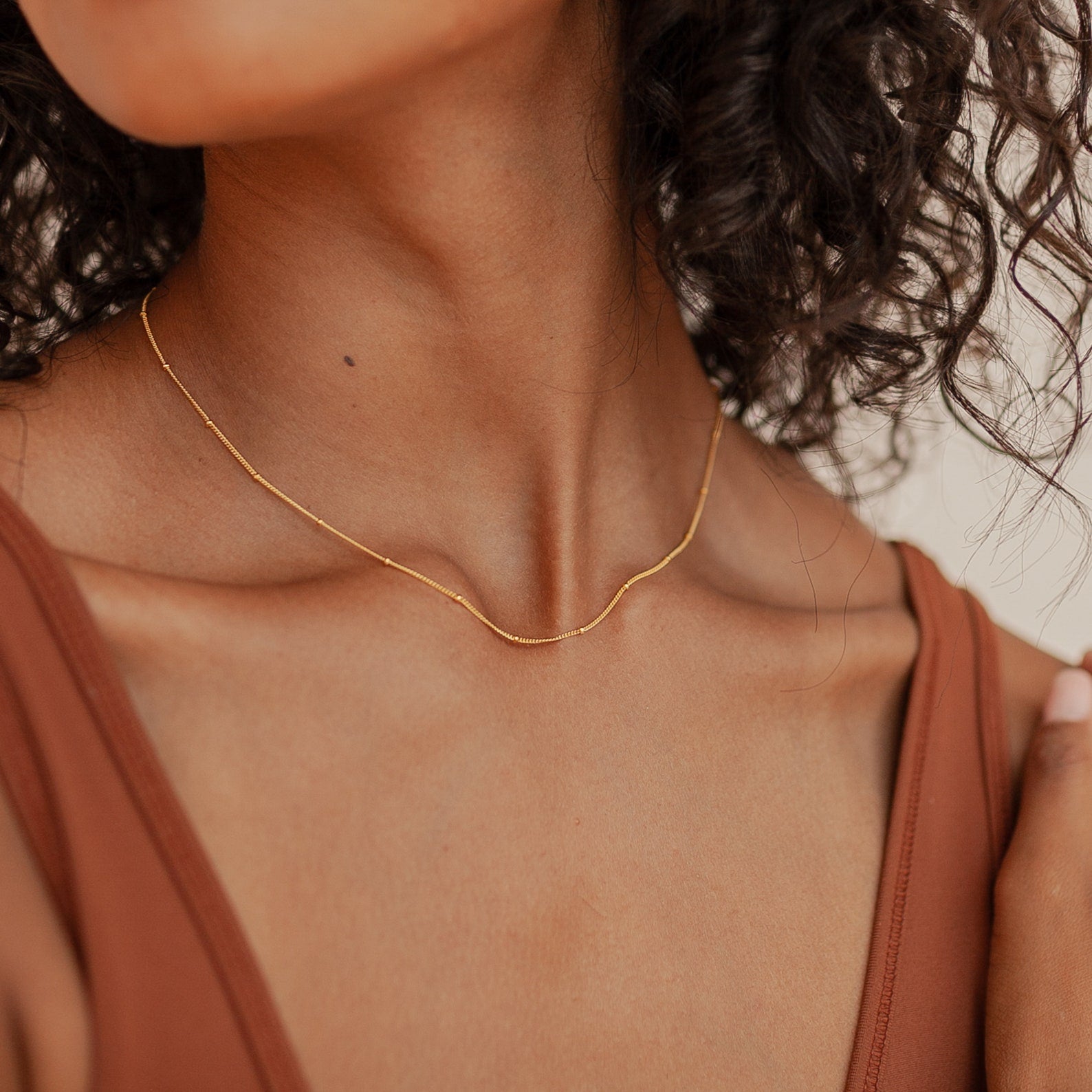 A close-up view of a woman's neck and collarbone showcases a delicate Satellite Necklace in gold. The fine chain is interspersed with tiny spherical beads spaced evenly along its length, catching light gently