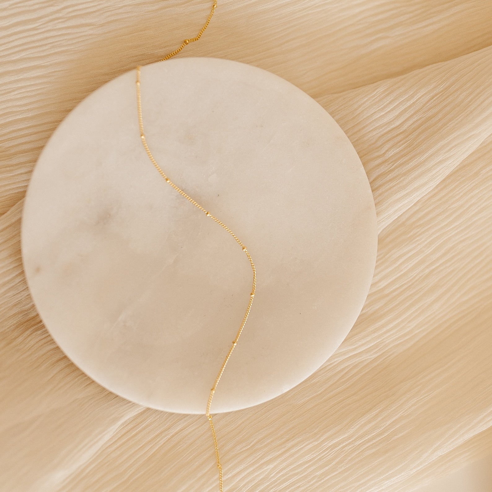 A Satellite Necklace laid across a round marble slab on a beige, crinkled fabric background.