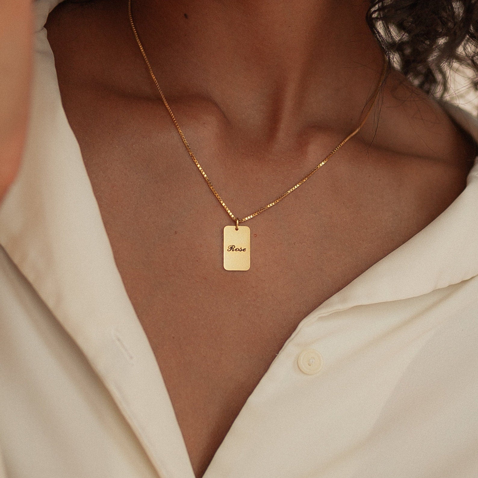 The Name Tag Necklace features a rectangular rose pendant, making it a perfect piece of personalized name jewelry, shown worn with a cream-colored shirt.