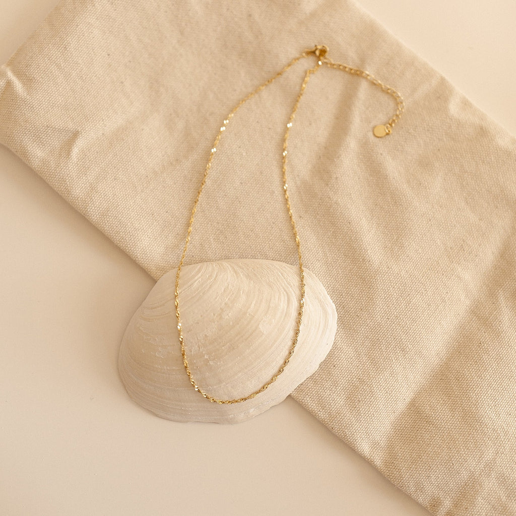 Singapore Twist Necklace resting in a curved shape atop a white shell and neutral fabric, styled in a coastal-inspired flat lay.