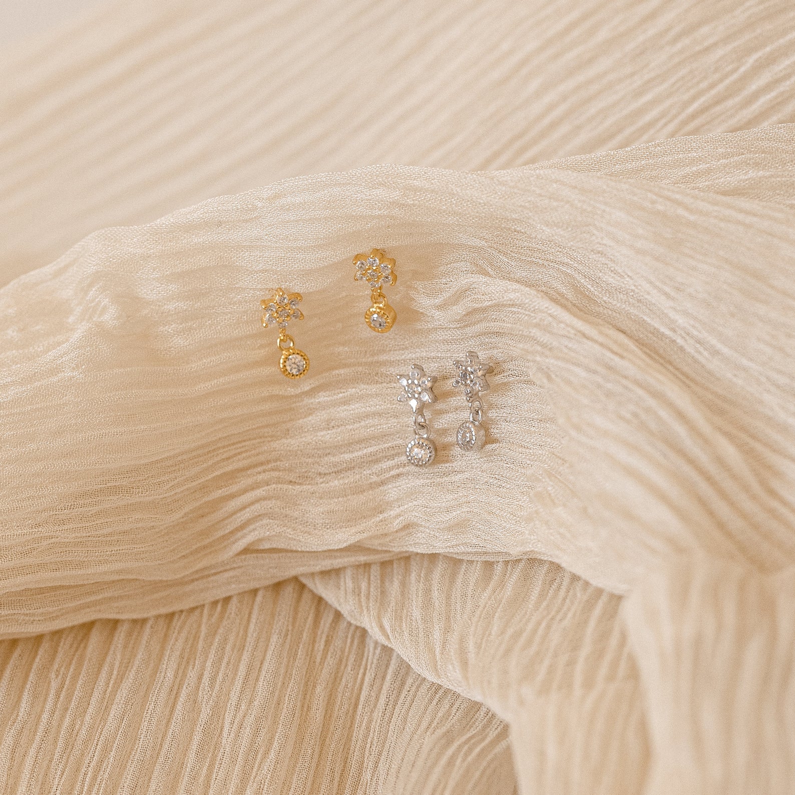 Two pairs of Lexi Flower Studs, featuring delicate gold and silver floral designs, are displayed on cream-colored textured fabric.