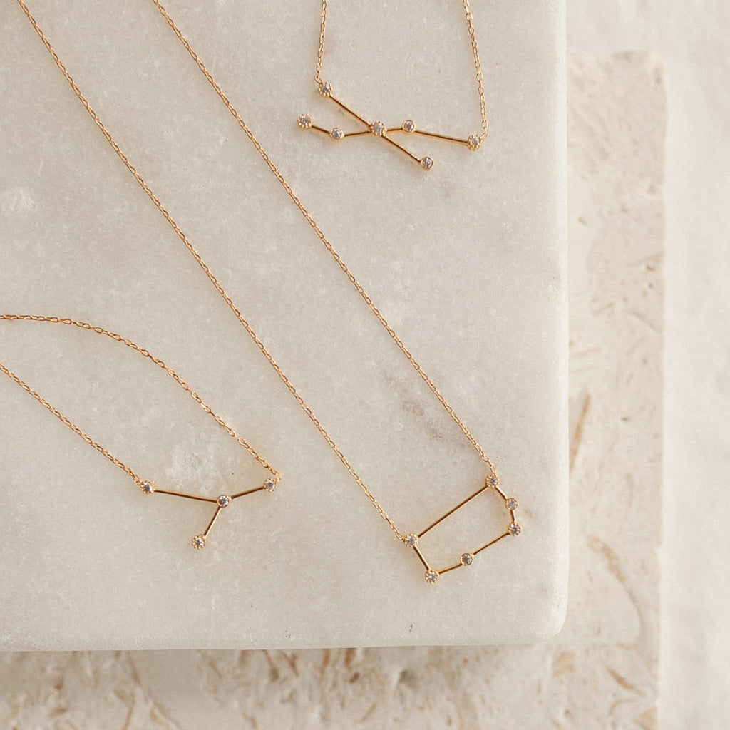 Three gold Constellation Necklaces, each inspired by the stars, are displayed on a light marble and textured stone surface.