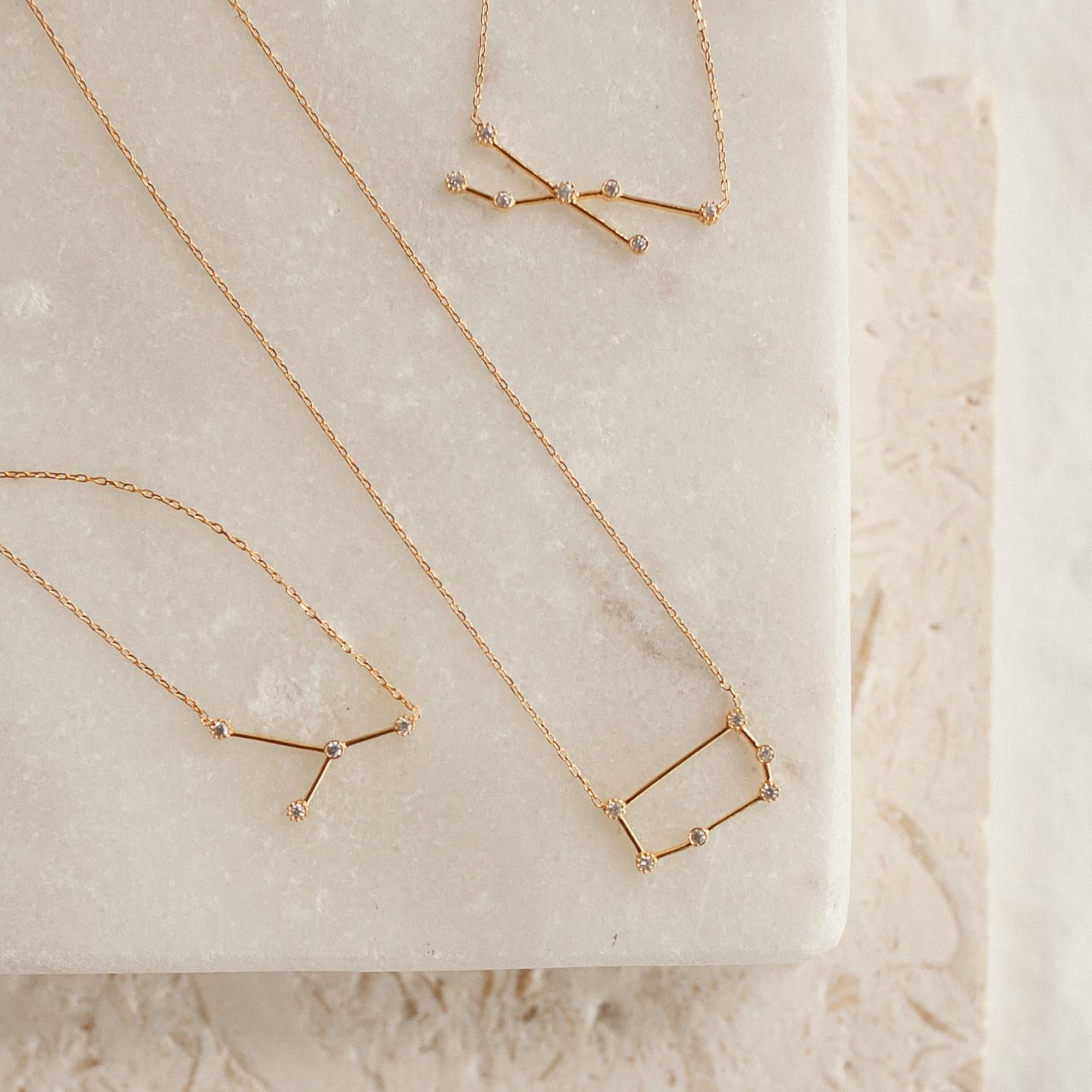 Three gold Constellation Necklaces, each inspired by the stars, are displayed on a light marble and textured stone surface.