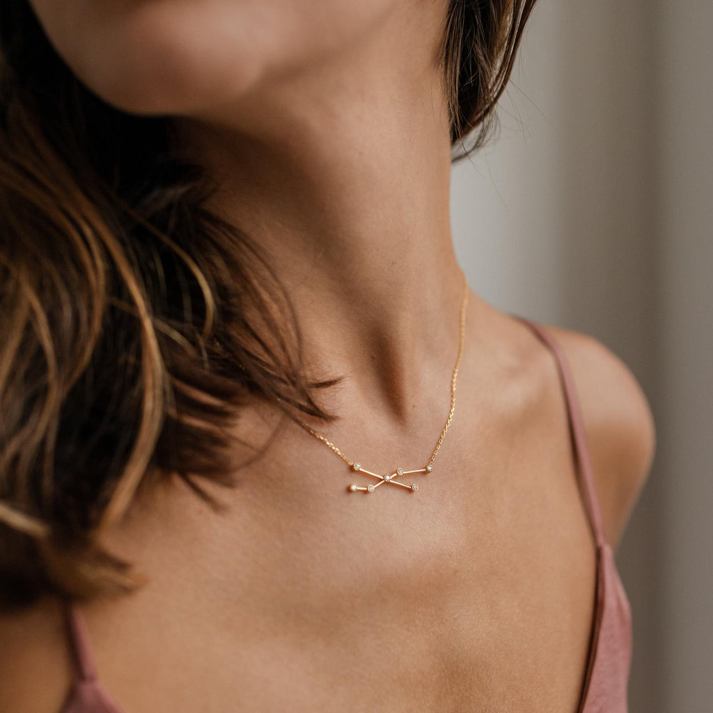 A woman in a pink spaghetti strap top wears the Constellation Necklace, featuring two intersecting gold arrows.