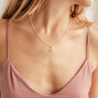 A woman wearing an Old English Initial Necklace with Curb Chain that has a gold M pendant and a mauve spaghetti strap top.