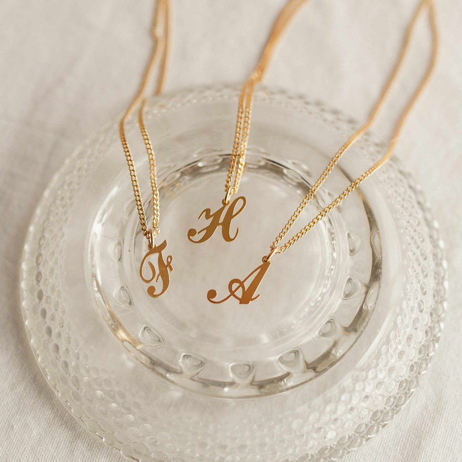 Three gold Old English Initial Necklaces with Curb Chains—featuring the letters F, H, and A—are displayed on a clear glass dish for a chic goth-glam look.