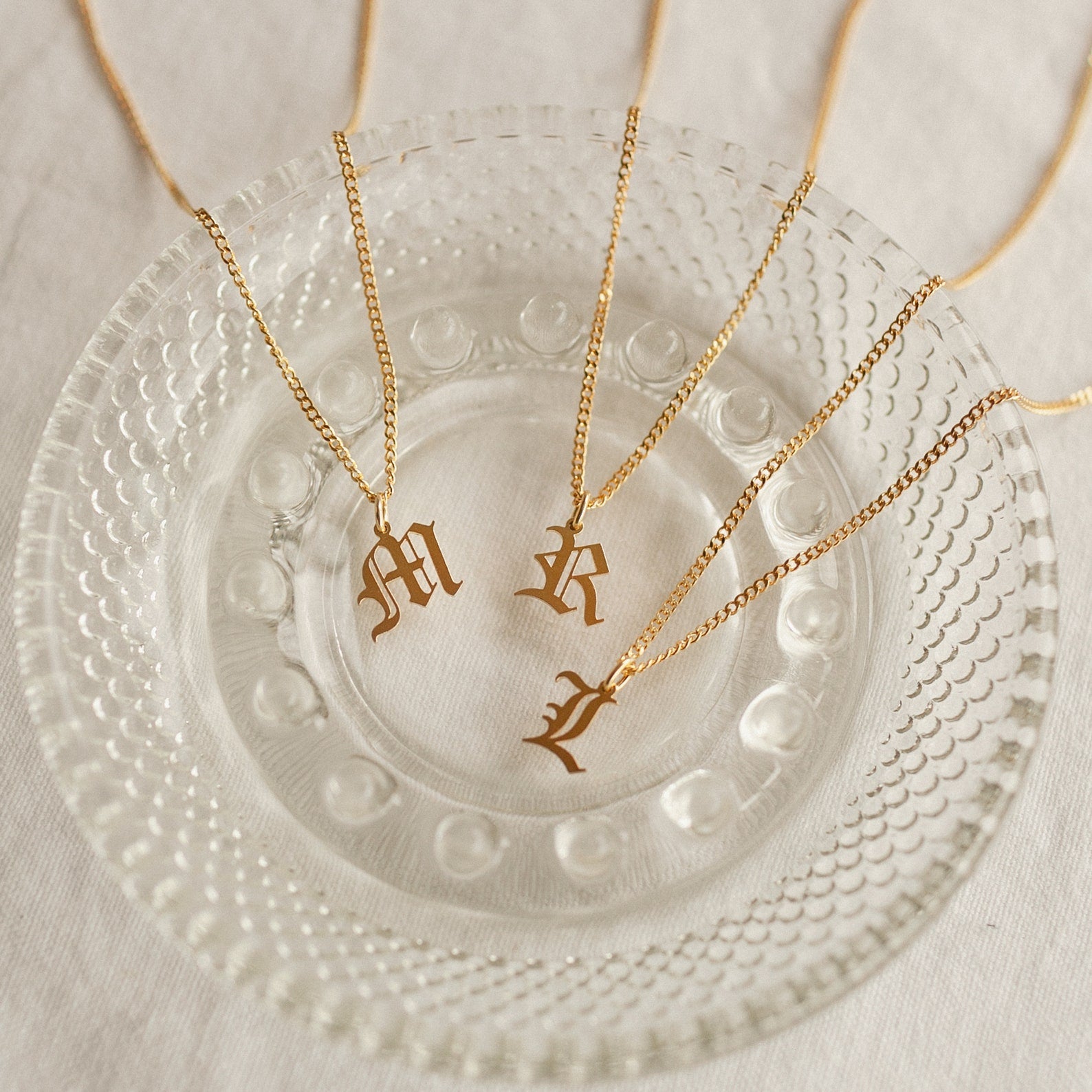 Three gold Old English Initial Necklace with Curb Chain pieces—featuring “M,” “R,” and “L” pendants—rest elegantly on a clear glass dish.