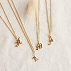 Four Old English Initial Necklaces with Curb Chain, featuring H, L, M, and A letter pendants in gold, are displayed on a light textured fabric for a stylish goth-glam look.