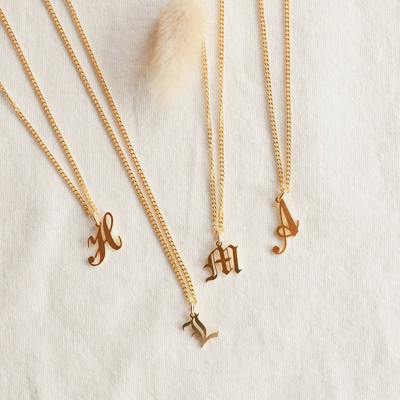 Four Old English Initial Necklaces with Curb Chain, featuring H, L, M, and A letter pendants in gold, are displayed on a light textured fabric for a stylish goth-glam look.