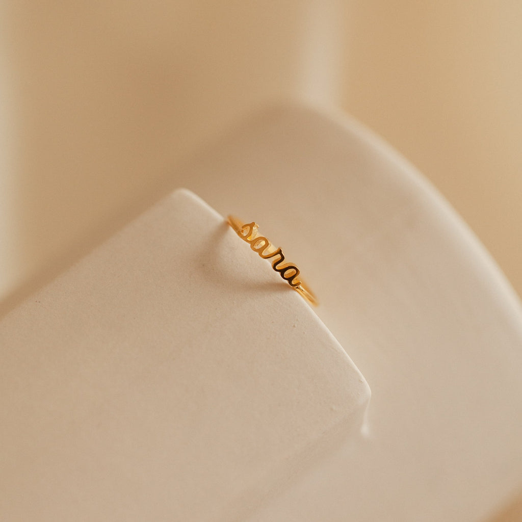 Petite Tiny Name Ring featuring the name "Sara" in cursive, displayed on a beige, curved surface. This delicate gold ring offers a personalized touch to your style.