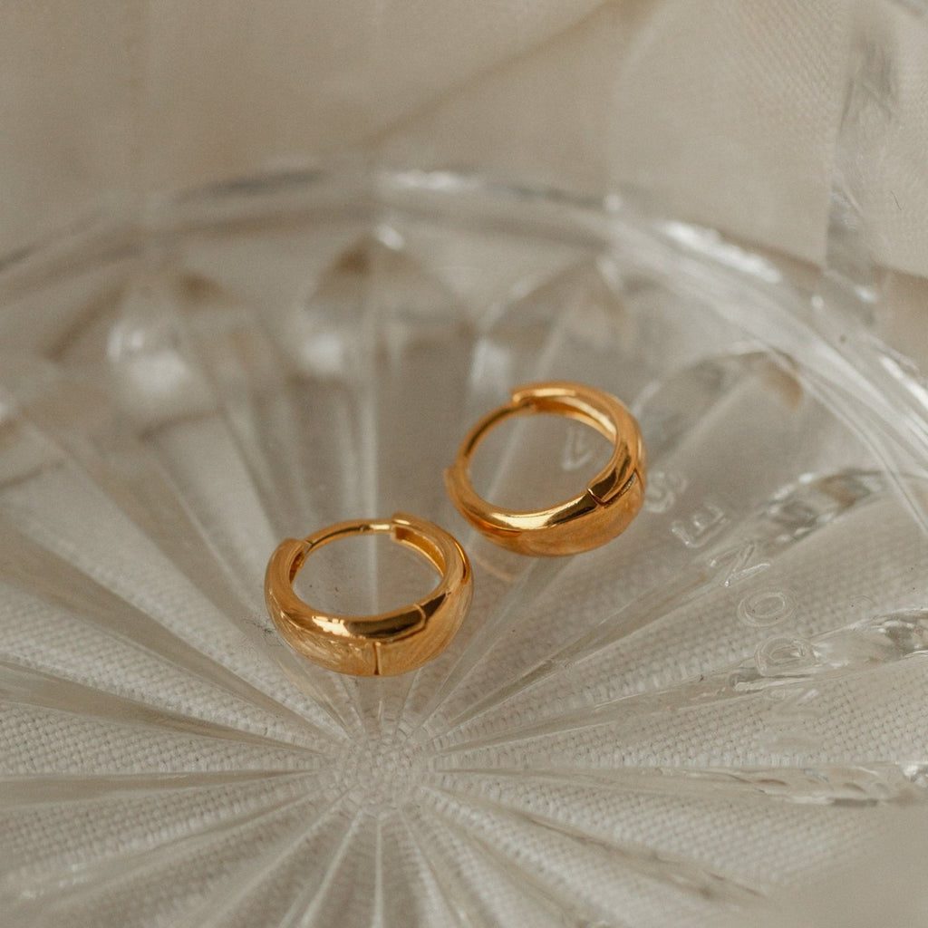 The Lucy Hinged Huggies, a pair of minimalist gold hoop earrings, are displayed on a clear textured glass dish against a beige fabric background.