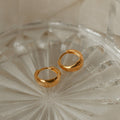 The Lucy Hinged Huggies, a pair of minimalist gold hoop earrings, are displayed on a clear textured glass dish against a beige fabric background.