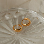 The Lucy Hinged Huggies, a pair of minimalist gold hoop earrings, are displayed on a clear textured glass dish against a beige fabric background.
