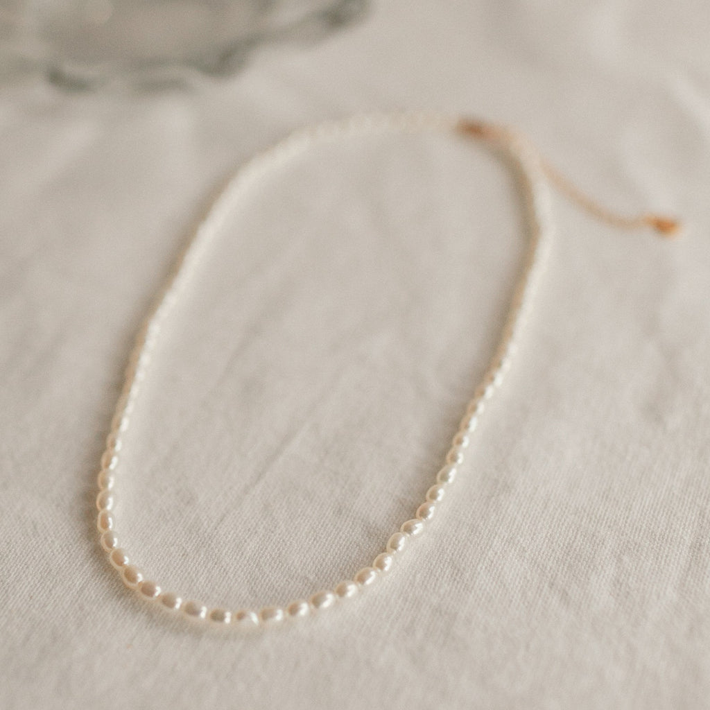 Delicate single strand pearl necklace with small, evenly spaced oval pearls, laid flat on a soft beige fabric surface.
