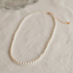 Delicate single strand pearl necklace with small, evenly spaced oval pearls, laid flat on a soft beige fabric surface.