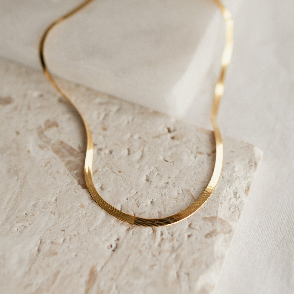A close-up image of a thick herringbone necklace in yellow gold resting in gentle curves on a textured beige stone surface. The necklace displays a fluid, flat silhouette with fine, closely linked metalwork that gives it a smooth and reflective finish.