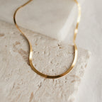 A close-up image of a thick herringbone necklace in yellow gold resting in gentle curves on a textured beige stone surface. The necklace displays a fluid, flat silhouette with fine, closely linked metalwork that gives it a smooth and reflective finish.