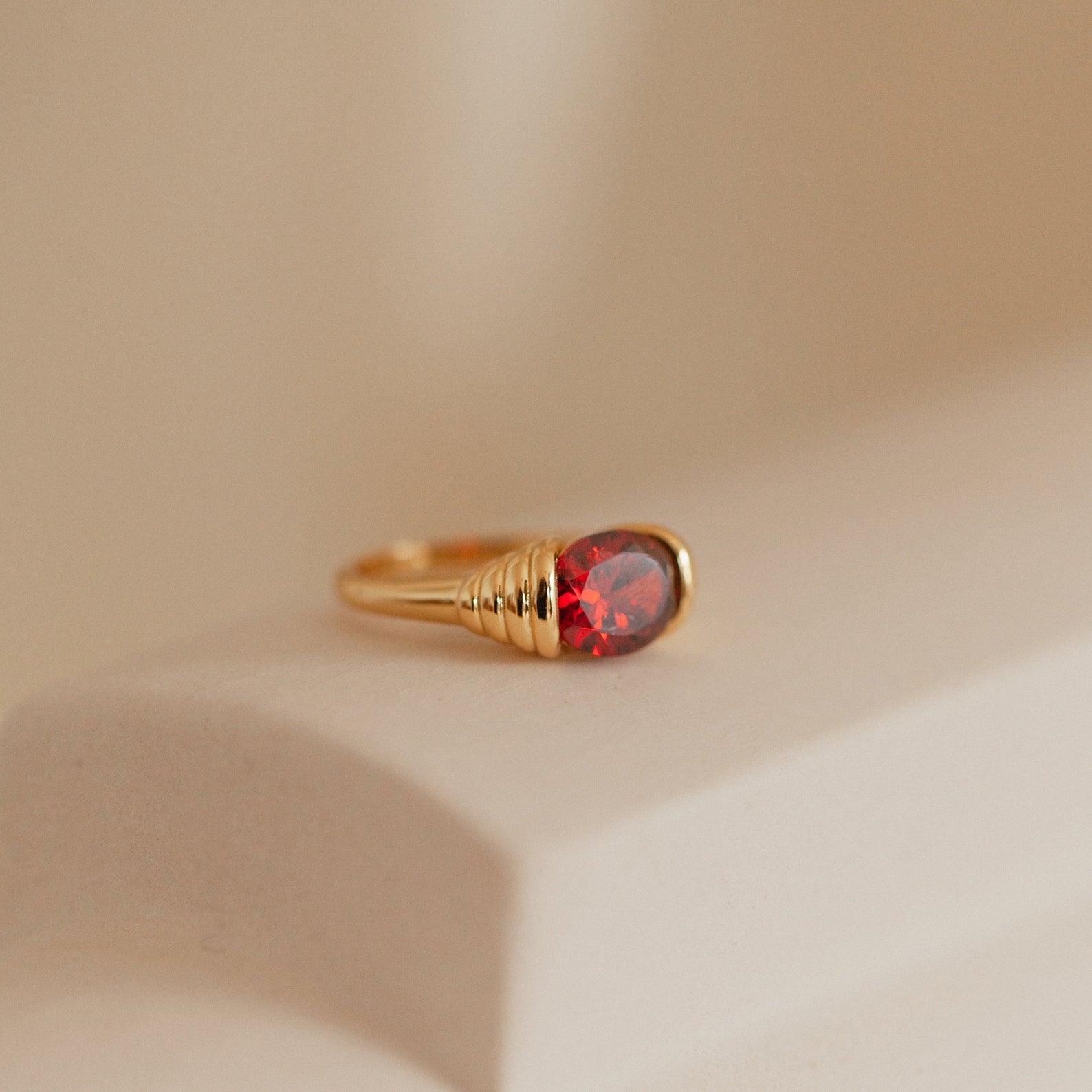 The Garnet Signet Ring features a vintage-inspired design and a striking red gemstone, displayed on a smooth cream surface.