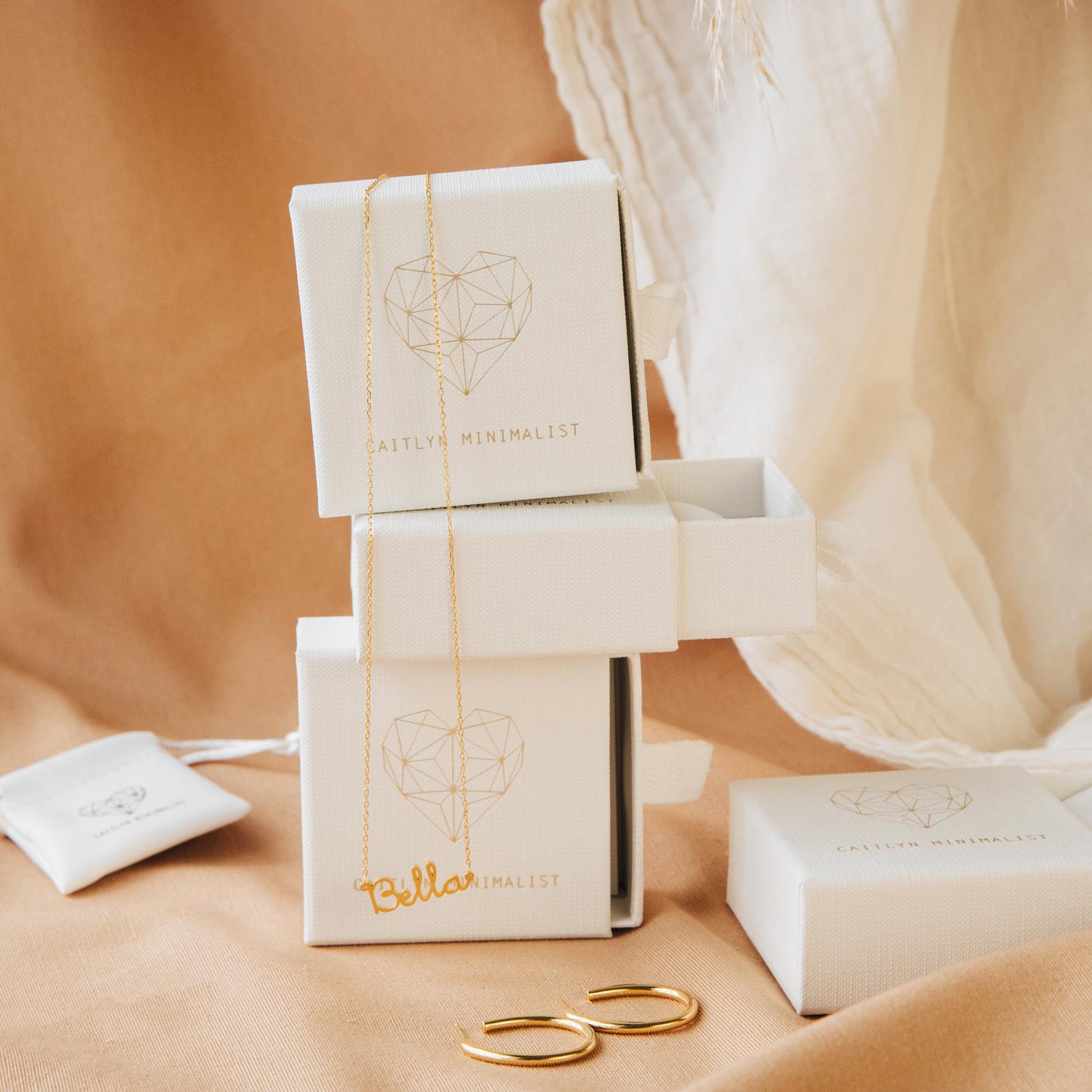 Dainty Name Necklace displayed with hoop earrings and white jewelry boxes on a soft beige fabric background—perfect for minimalist jewelry lovers.