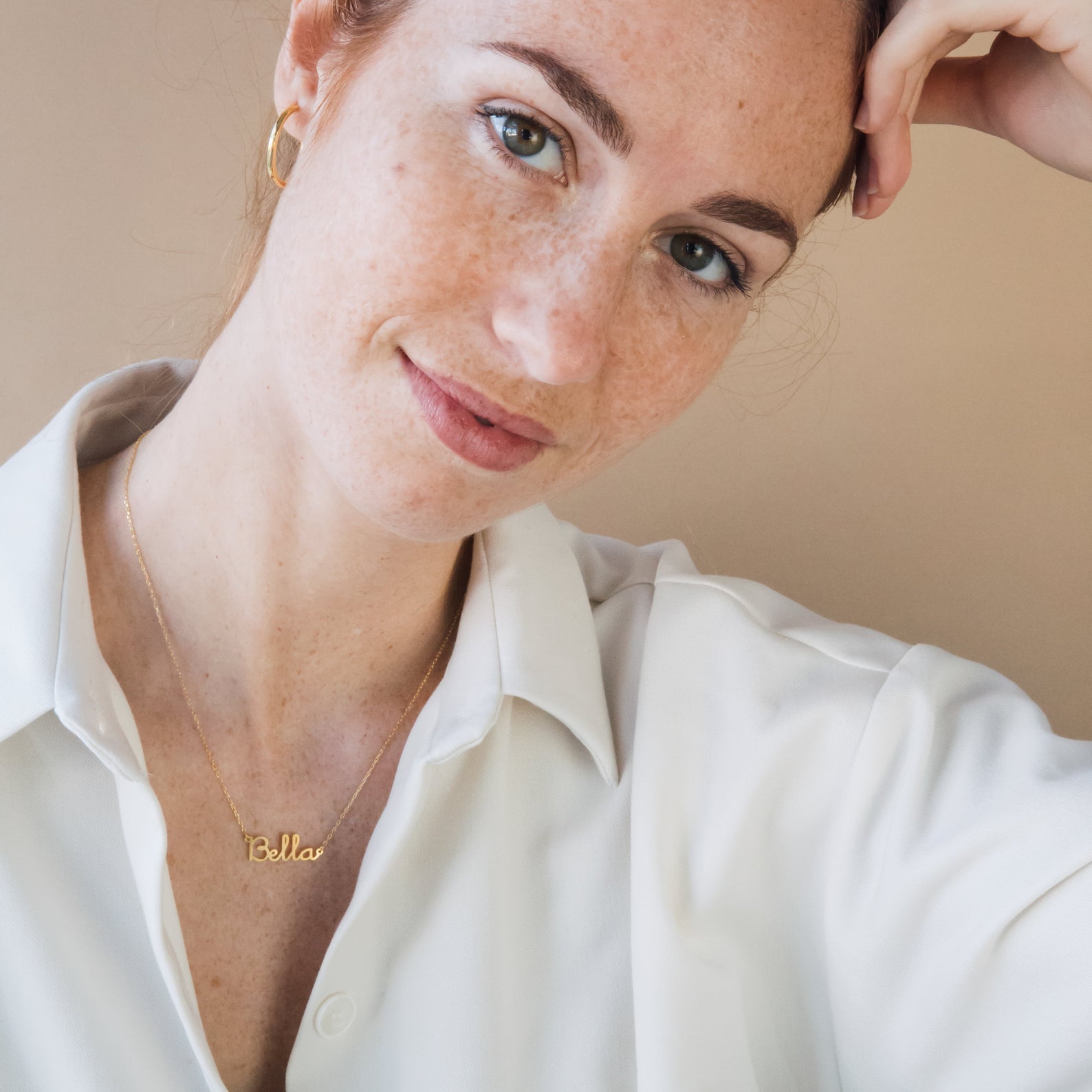 A red-haired, freckled woman wearing a white shirt gently smiles at the camera, elegantly showcasing the Dainty Name Necklace and her love for minimalist jewelry.