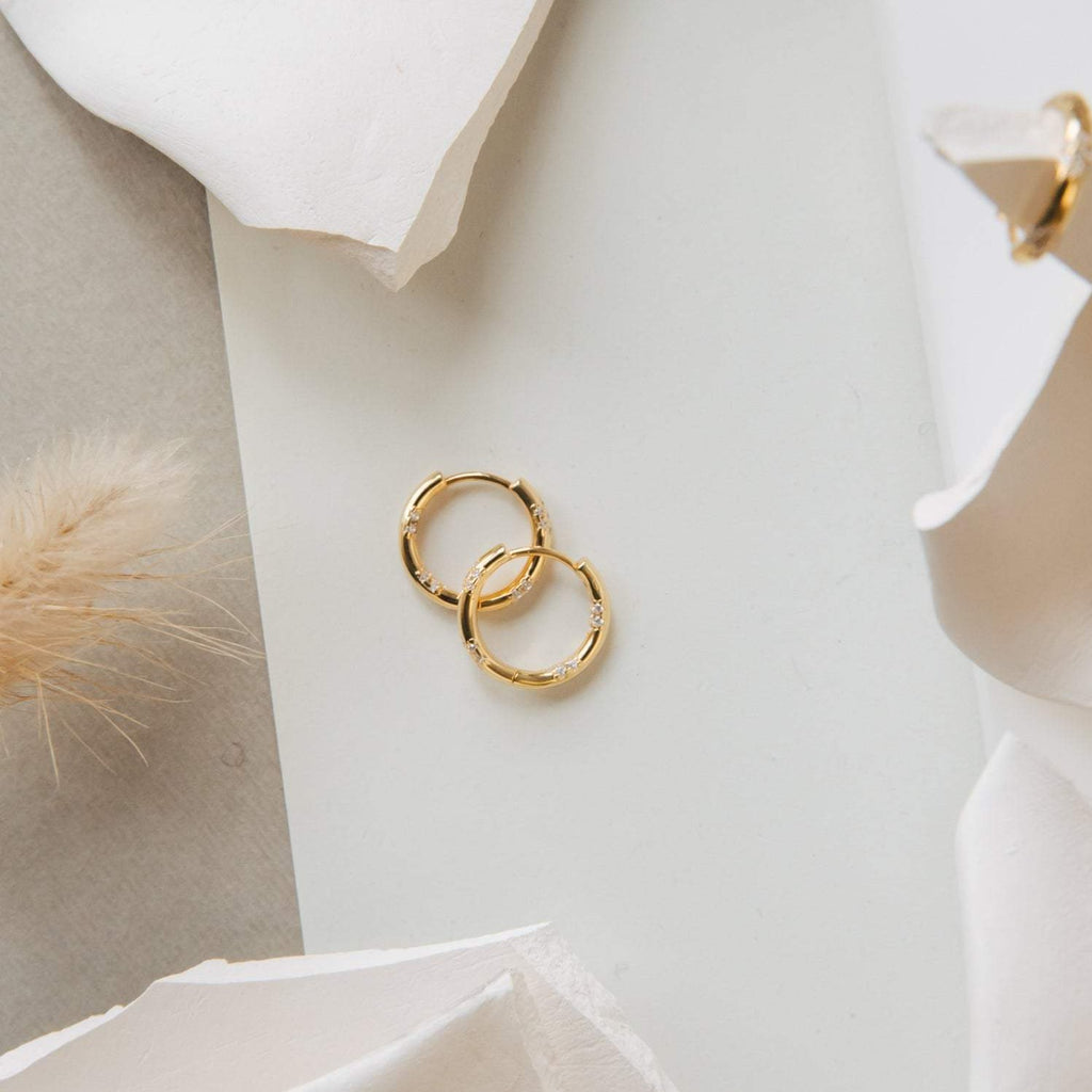 Two Kaia Diamond Huggies rest on a white surface, accented by abstract white shapes and nearby beige decorations.