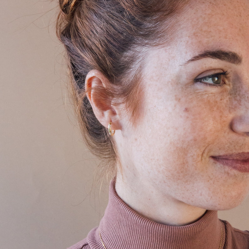 Red-haired woman with a bun, wearing a pink turtleneck and Kaia Diamond Huggies, smiles softly.