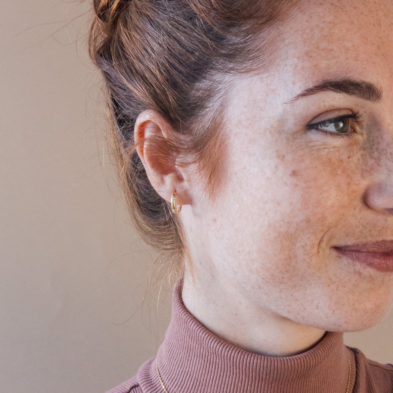 Red-haired woman with a bun, wearing a pink turtleneck and Kaia Diamond Huggies, smiles softly.