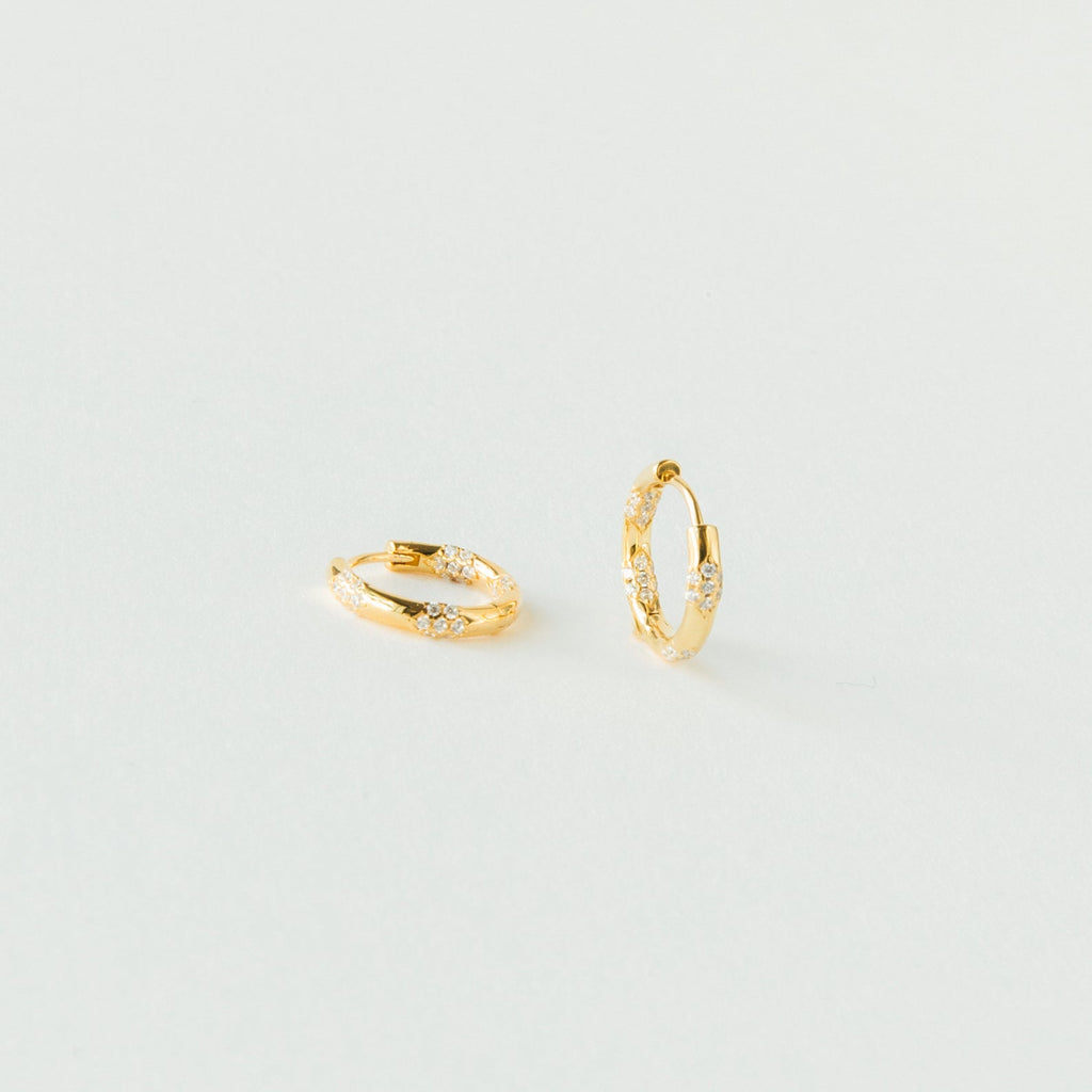The Kaia Diamond Huggies are small gold hoop earrings with clear stones, adding subtle sparkle. Their dainty diamond design is beautifully showcased against a plain white background.