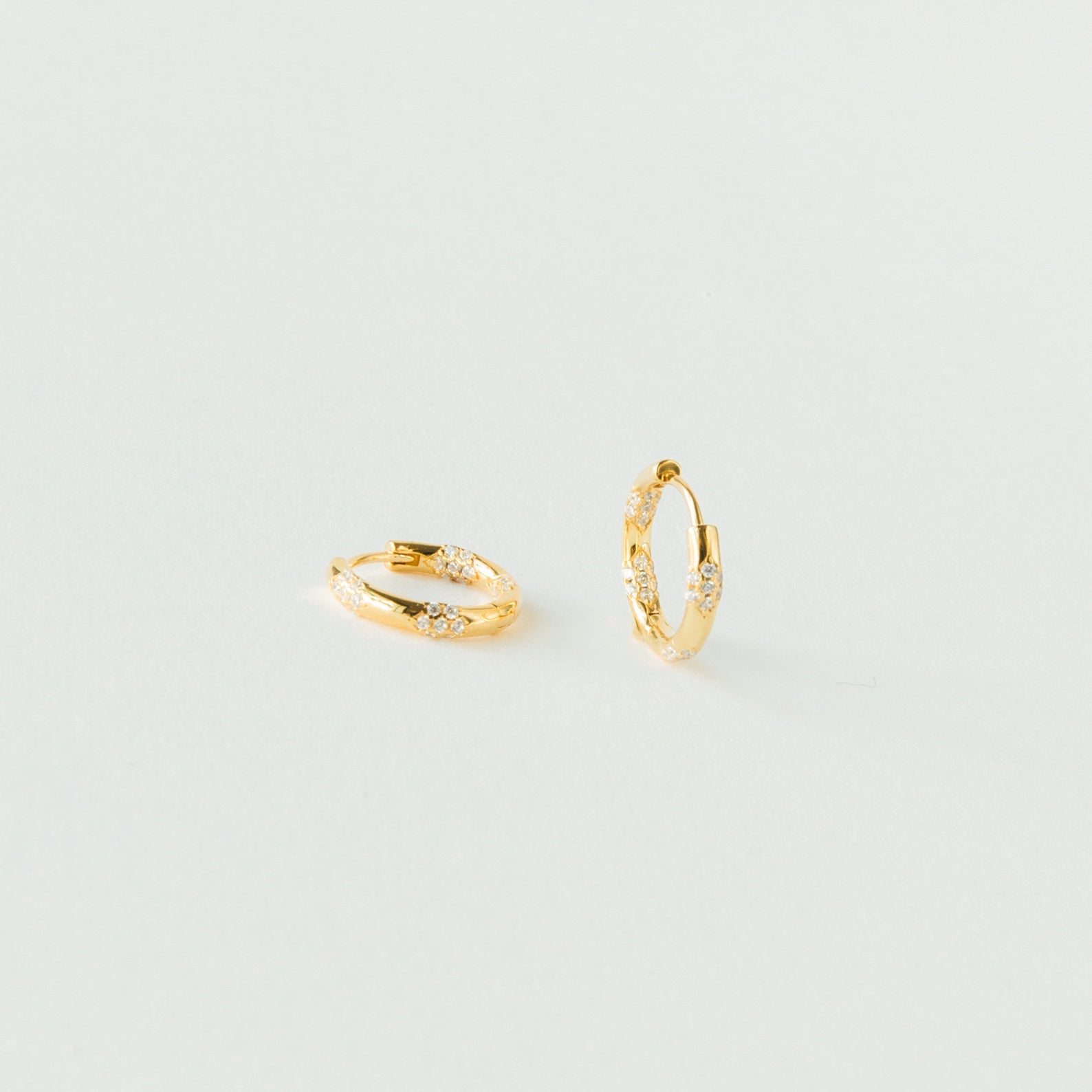 The Kaia Diamond Huggies are small gold hoop earrings with clear stones, adding subtle sparkle. Their dainty diamond design is beautifully showcased against a plain white background.