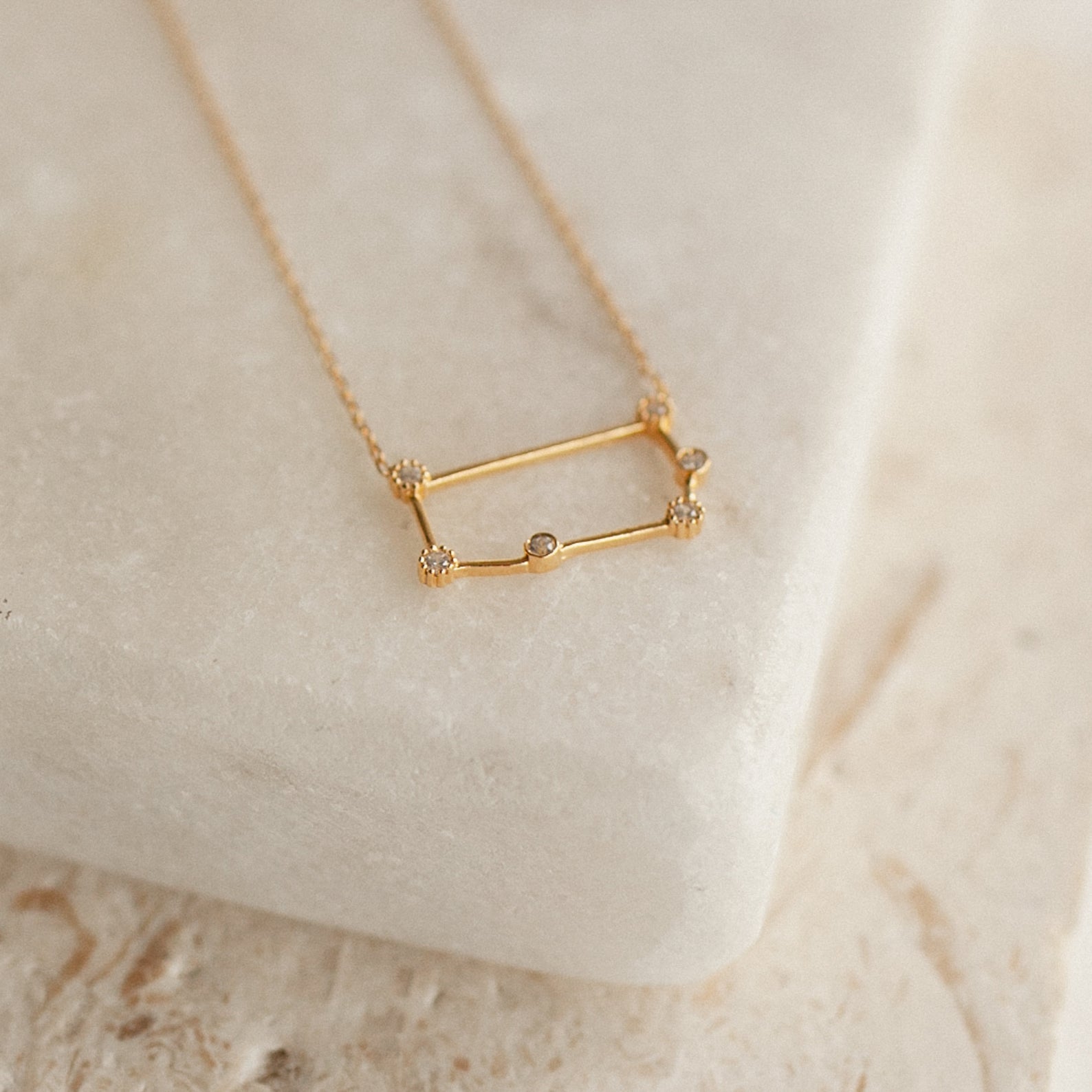 Constellation Necklace with a rectangular pendant set with small gemstones, showcased on a smooth, light-colored stone surface for a celestial look.