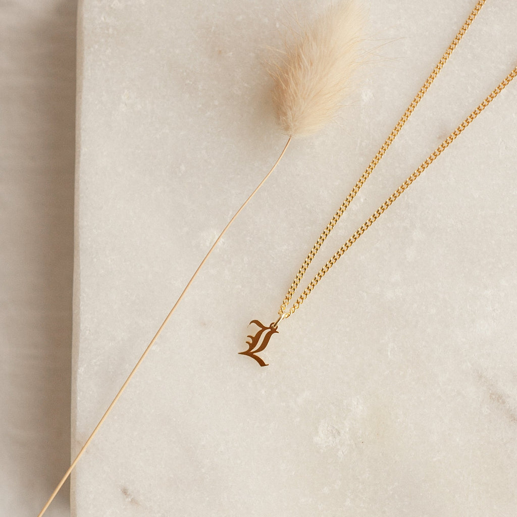 The Old English Initial Necklace with Curb Chain features an L pendant on a marble surface beside a fluffy beige dried plant, bringing a goth-glam style to your look.