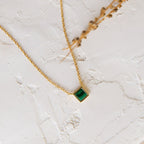 Gold necklace with a square bezel set green gemstone pendant resting on a textured white surface next to a dried botanical stem.